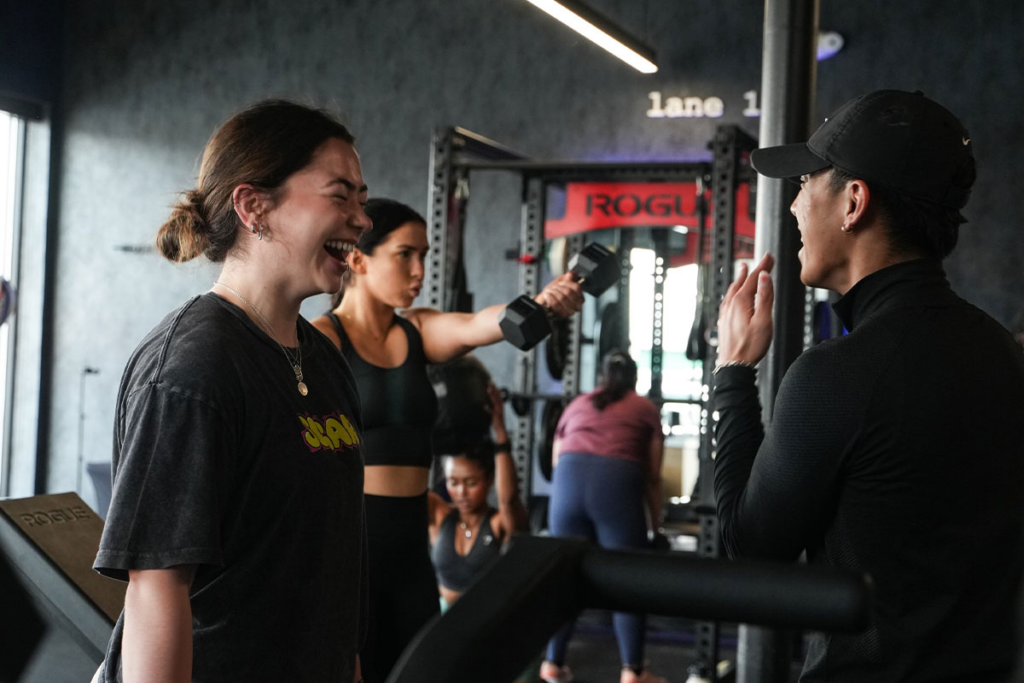 A personal trainer coaching a female client in a group fitness class. The client is laughing with the trainer.