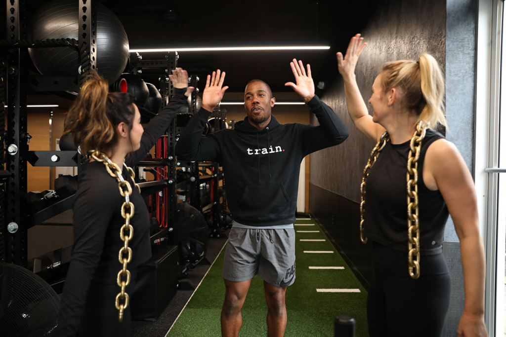A personal trainer giving a high five to two female clients with gold weight chains on their neck.